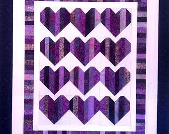 May include: A handmade quilt with a repeating heart design in various shades of purple. The hearts are made from vertical fabric strips, framed by a white border and a striped purple border. The quilt is set against a dark blue background.