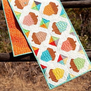 May include: A quilted table runner with a pattern of colourful acorns and kites on a white background. The runner has a turquoise border and an orange backing. Finished size is 38cm x 165cm.