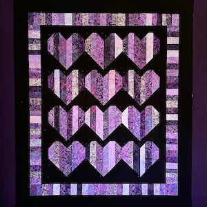 May include: A handmade quilt featuring a pattern of purple hearts against a black background. The hearts are constructed from various patterned fabrics in shades of purple, lavender, and white. The quilt has a black border and a striped outer edge.