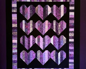 May include: A patchwork quilt with a design of purple hearts on a black field. The hearts are made from strips of patterned fabric in purple, lilac, and white. The quilt has a black border and a striped outer edge.