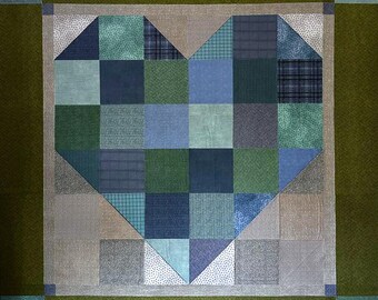 May include: A square quilt with a heart design. The heart is made from blue and green fabric squares, with a neutral border and a green background. The quilt is a patchwork design, suitable for a bed or wall hanging.