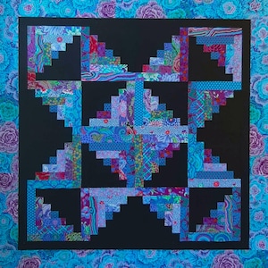 May include: A handmade quilt with a central black square featuring a geometric design in shades of blue, purple, and teal. The quilt's border is a floral pattern in similar colors. The quilt is square in shape.