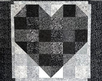 May include: A handmade quilt with a heart design. The heart is made of black, white, and grey patterned squares. The quilt has a white border and a black floral background, creating a striking contrast.