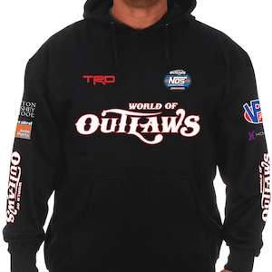 Pode incluir: Camisola preta com capuz com o texto "WORLD OF OUTLAWS" em branco e vermelho. A camisola tamb&eacute;m apresenta os log&oacute;tipos "TRD" e "NOS" e outros log&oacute;tipos nas mangas. A pessoa na imagem est&aacute; a usar um bon&eacute; preto e cal&ccedil;as de ganga azuis.