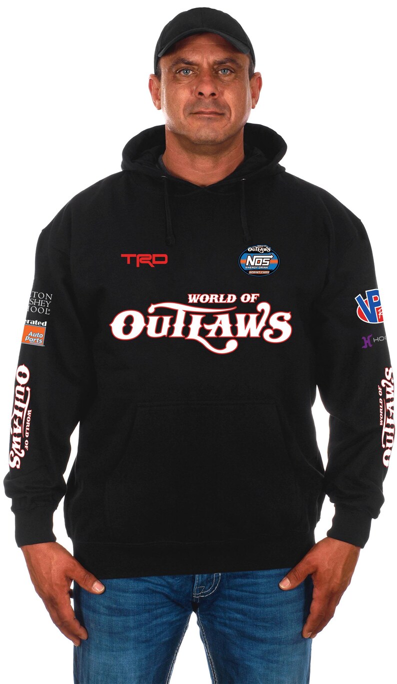 Pode incluir: Camisola preta com capuz com o texto "WORLD OF OUTLAWS" em branco e vermelho. A camisola tamb&eacute;m apresenta os log&oacute;tipos "TRD" e "NOS" e outros log&oacute;tipos nas mangas. A pessoa na imagem est&aacute; a usar um bon&eacute; preto e cal&ccedil;as de ganga azuis.