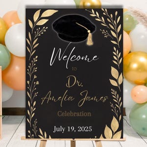 May include: A black graduation welcome sign with gold accents. The sign features a graduation cap illustration and the words "Welcome to Dr. Amelia James Celebration July 19, 2025". The sign is on a wooden easel.