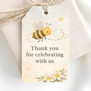 以下が含まれることがあります： 漫画のミツバチと「Thank you for celebrating with us」の文字が描かれた水彩デザインのギフトタグ。タグはクリーム色の背景に花と星のアクセントがあり、紐で結ばれています。