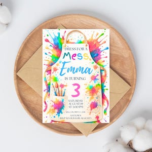 Puede incluir: Una invitaci&oacute;n de cumplea&ntilde;os con un dise&ntilde;o de delantal de acuarela y salpicaduras de pintura coloridas. La invitaci&oacute;n dice "Dress for a Mess Emma is turning 3" con detalles del evento. La invitaci&oacute;n est&aacute; en un plato de madera con un sobre.