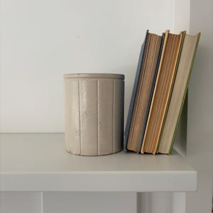 May include: A white ceramic cylindrical container with vertical lines sits on a white shelf next to a stack of three books. The books are bound in blue, yellow, and green.