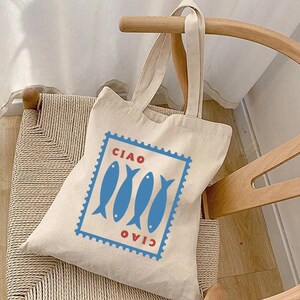 May include: A beige canvas tote bag featuring a blue and red design of three fish and the word "CIAO" above and below. The bag has two handles and is resting on a wooden chair with a woven seat.