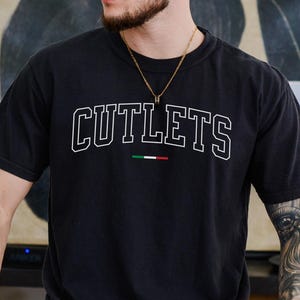 May include: Black t-shirt with the word "CUTLETS" in white, outlined letters. Below the word is a small Italian flag. The shirt is short-sleeved and appears to be made of a soft material.