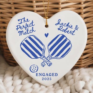 May include: A heart-shaped ceramic ornament with blue pickleball paddles and the text "The Perfect Match" and "Jackie & Robert." The ornament also includes the word "Engaged" and the year "2025."