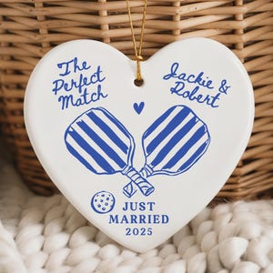 May include: A heart-shaped ceramic ornament with blue pickleball paddles and text that reads "The Perfect Match" and "Jackie & Robert Just Married 2025". The ornament hangs from a gold string.
