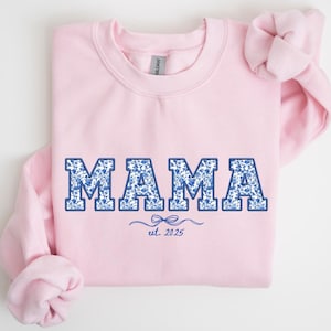 May include: A light pink sweatshirt with the word "MAMA" in blue floral print letters. Below the word "MAMA" is the text "est. 2025" in a dark blue script font. The sleeves are rolled up.
