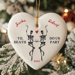 May include: Heart-shaped white ceramic ornament with gold string, featuring skeleton couple design. Text includes "Jackie" and "Robert", "'Til Death Do Us Part", and the year "2025". A festive decoration for a wedding or anniversary.