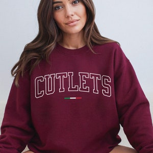 May include: A maroon sweatshirt with the word "CUTLETS" in white, outlined letters. Below the word is a small Italian flag. The sweatshirt is a crew neck style.