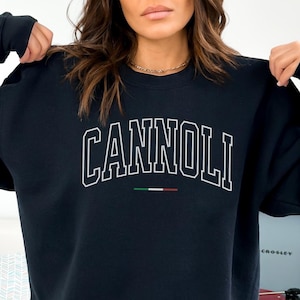 Może przedstawiać: Granatowa bluza z napisem "CANNOLI" w białych, łukowatych literach. Poniżej słowa znajduje się mały poziomy pasek w kolorach włoskiej flagi. Bluza ma okrągły dekolt.