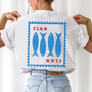 May include: White t-shirt with a blue and red graphic. The graphic features four blue fish inside a blue postage stamp design, with the words "CIAO" and "OVIO" in red. The shirt is made of a soft material.