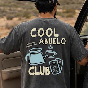 May include: A charcoal gray short-sleeve t-shirt featuring a back graphic with the text COOL ABUELO CLUB in cream. The design includes a light blue coffee carafe, a steaming cup of coffee, and two dominoes, with the text powered by cafecito & nap.