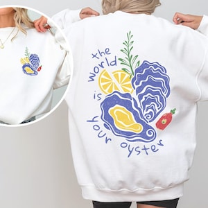 Puede incluir: Sudadera blanca con un gráfico de una ostra, un limón, romero y salsa picante. El texto "the world is your oyster" está impreso en la sudadera.