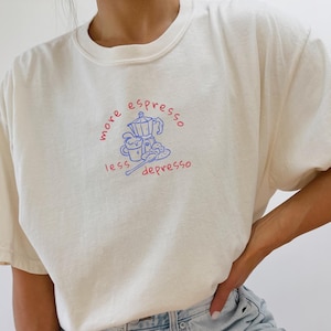 Puede incluir: Una camiseta blanca con un gráfico azul y rojo que dice "more espresso less depresso" con un dibujo de una cafetera y tazas de café.