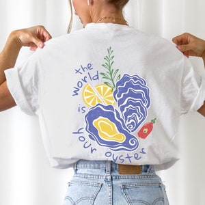 May include: White t-shirt with a graphic design of oysters, lemon, rosemary, and a red hot sauce bottle. The text "the world is your oyster" is printed on the shirt.
