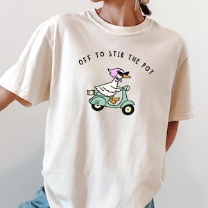 May include: Cream-colored t-shirt with the phrase "OFF TO STIR THE POT" printed above a cartoon duck riding a teal scooter. The duck wears a pink headscarf and sunglasses. The scooter has a red luggage rack.