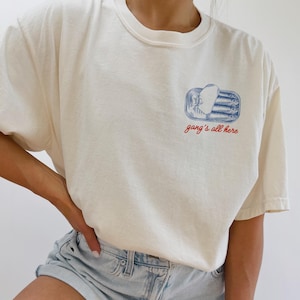 May include: Cream-colored t-shirt with a small graphic of a sardine can in blue and white, with the text "gang's all here" in red. The shirt has a relaxed fit and short sleeves. The model is wearing light blue denim shorts.