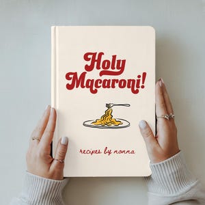 May include: A cream-coloured cookbook titled "Holy Macaroni!" with red lettering and a pasta illustration. The words "recipes by nonna" are printed below the illustration. The book is held by two hands.