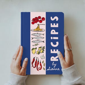 May include: A blue recipe book with the word "RECIPES" in white, and illustrations of food. The book has a pink vertical stripe with food illustrations. The text "by Sienna" is written in cursive. The book is held by a person.