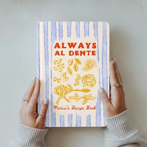 May include: A recipe book with a cream cover and blue vertical stripes, held by hands. The cover reads "ALWAYS AL DENTE" with pasta illustrations in yellow and "Maria's Recipe Book" in red.