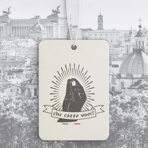 May include: A cream-colored tag with a black graphic of a hand gesture and the text "che cazzo vuoi?" in a banner. The tag has a white string and a backdrop of a cityscape.