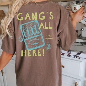 May include: A brown short-sleeve t-shirt featuring a graphic on the back. The design includes a light blue tin of sardines with the text "SARDINES WILD CAUGHT IN OLIVE OIL" and the phrases "GANG'S ALL HERE!" and "better together" in lime green.