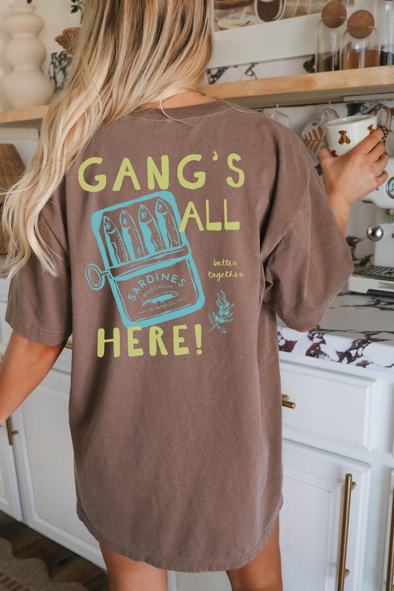 May include: A brown short-sleeve t-shirt featuring a graphic on the back. The design includes a light blue tin of sardines with the text "SARDINES WILD CAUGHT IN OLIVE OIL" and the phrases "GANG'S ALL HERE!" and "better together" in lime green.