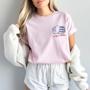 May include: A light pink short-sleeve t-shirt featuring a blue graphic of an open tin of sardines on the left chest. Below the graphic, the text "gang's all here" is printed in a red cursive font. The shirt has a crew neckline and a relaxed fit.