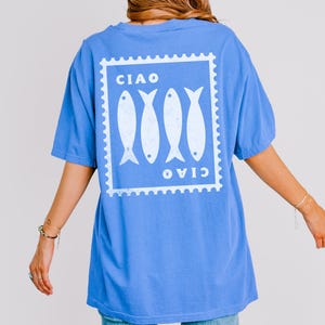 May include: A blue t-shirt with a white graphic on the back featuring four fish inside a postage stamp design. The word "CIAO" is printed above and below the fish. The shirt has short sleeves and a relaxed fit.