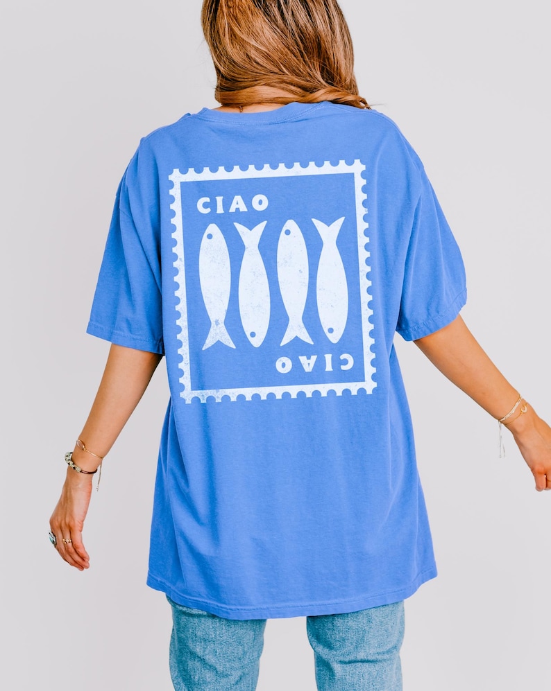 May include: A blue t-shirt with a white graphic on the back featuring four fish inside a postage stamp design. The word "CIAO" is printed above and below the fish. The shirt has short sleeves and a relaxed fit.
