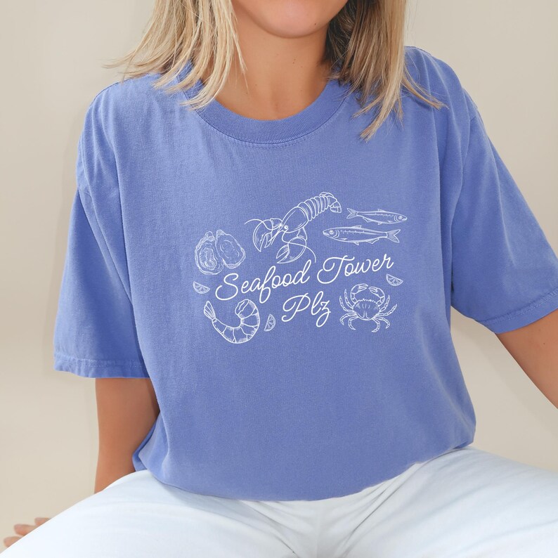 Peut inclure: Un t-shirt bleu clair avec un graphique blanc d'une tour de fruits de mer, comprenant un homard, des crevettes, des palourdes, un crabe et du poisson. Le texte "Seafood Tower Plz" est &eacute;crit en cursive.