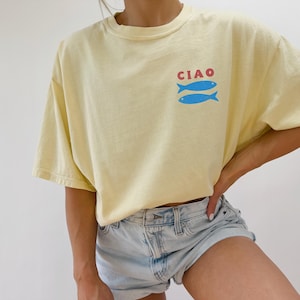 May include: A light yellow t-shirt with the word "CIAO" in red above two blue fish. The shirt has short sleeves and a relaxed fit. The model is wearing light blue denim shorts.