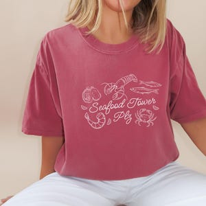 Peut inclure: Un t-shirt marron avec un motif blanc de fruits de mer. Le motif comprend un homard, des hu&icirc;tres, des poissons et un crabe. Le texte sur le t-shirt est "Seafood Tower Plz".