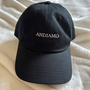 Pu&ograve; includere: Un cappello da baseball nero con la parola "ANDIAMO" ricamata in bianco sul davanti.