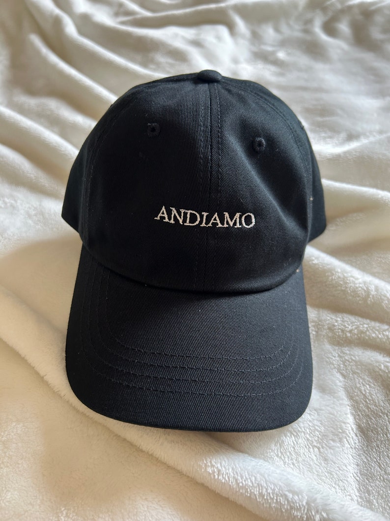 Pu&ograve; includere: Un cappello da baseball nero con la parola "ANDIAMO" ricamata in bianco sul davanti.