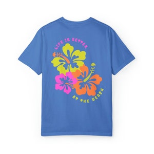 May include: A blue t-shirt with a floral design featuring yellow, orange, and pink hibiscus flowers. The text "LIFE IS BETTER AT THE BEACH" is printed above and below the flowers in a pink and yellow font.