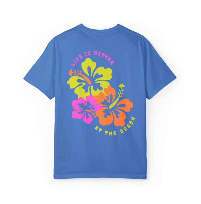 May include: A blue t-shirt with a floral design featuring yellow, orange, and pink hibiscus flowers. The text "LIFE IS BETTER AT THE BEACH" is printed above and below the flowers in a pink and yellow font.