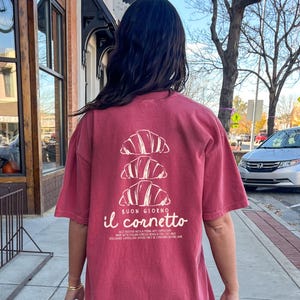 Op de afbeelding: Een rood T-shirt met een witte afbeelding van drie croissants die op elkaar gestapeld zijn. De tekst "BUON GIORNO il cornetto" is onder de croissants gedrukt. De tekst onder de afbeelding luidt "BEST ENJOYED WITH A FRING HOT CAPPUCCINO MADE WITH ITALIAN ESPRESSO BEANS & TILT MILK DISCLAIMER CAPPUCCINO SHOULD ONLY BE CONSUMED BEFORE 11AM"
