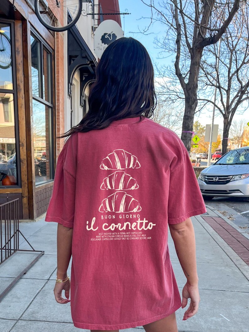 Op de afbeelding: Een rood T-shirt met een witte afbeelding van drie croissants die op elkaar gestapeld zijn. De tekst "BUON GIORNO il cornetto" is onder de croissants gedrukt. De tekst onder de afbeelding luidt "BEST ENJOYED WITH A FRING HOT CAPPUCCINO MADE WITH ITALIAN ESPRESSO BEANS & TILT MILK DISCLAIMER CAPPUCCINO SHOULD ONLY BE CONSUMED BEFORE 11AM"