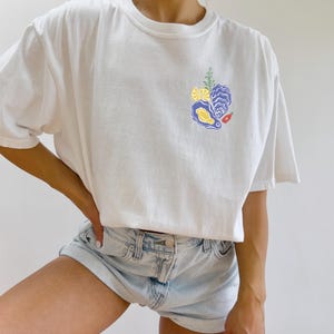 May include: White t-shirt with a graphic of an oyster, lemon, and a sprig of rosemary. The graphic is in shades of blue, yellow, and red.