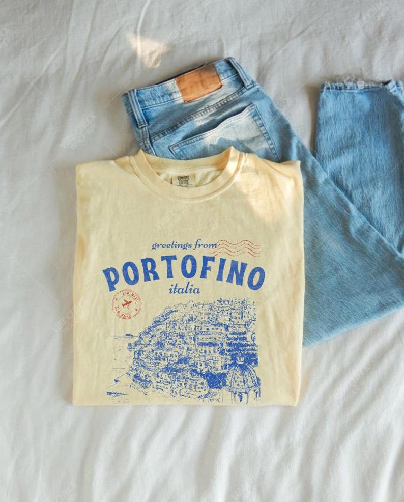 Może przedstawiać: Ż&oacute;łta koszulka z niebieskim nadrukiem przedstawiającym włoskie miasto Portofino. Nadruk zawiera tekst "greetings from Portofino italia".