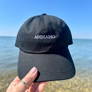 Pu&ograve; includere: Un cappello da baseball nero con un logo ricamato bianco che dice "ANDIAMO".