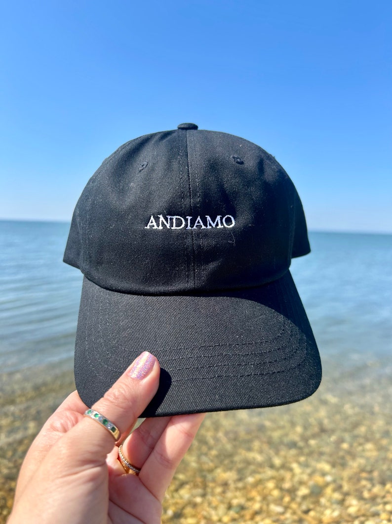 Pu&ograve; includere: Un cappello da baseball nero con un logo ricamato bianco che dice "ANDIAMO".